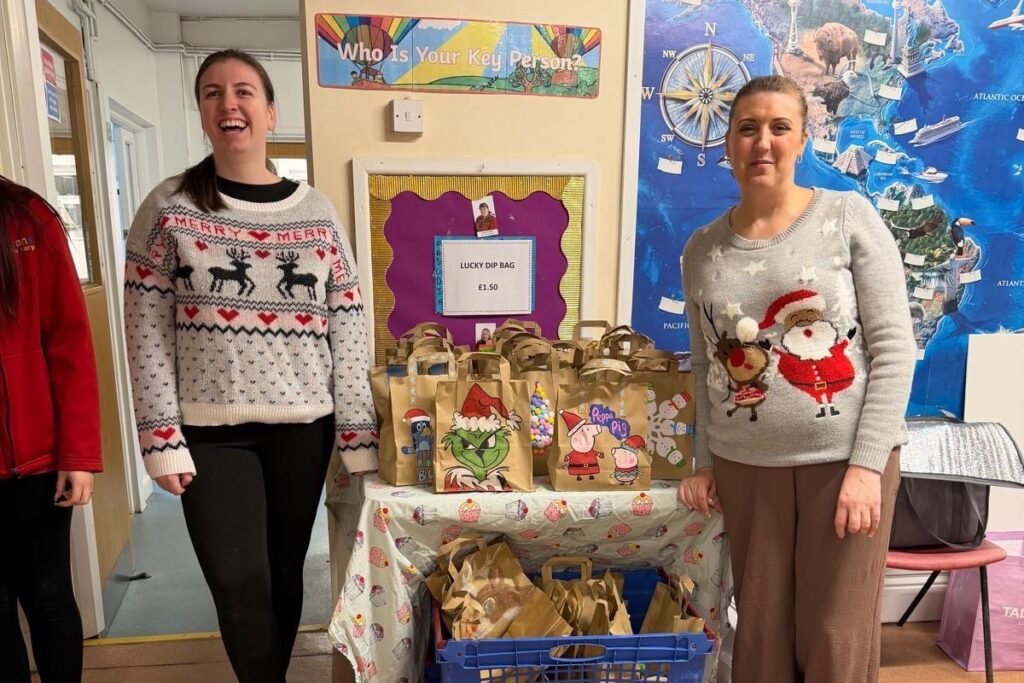 Two Rowans Day Nursery team members in Christmas jumpers beside a table of lucky dip bags
